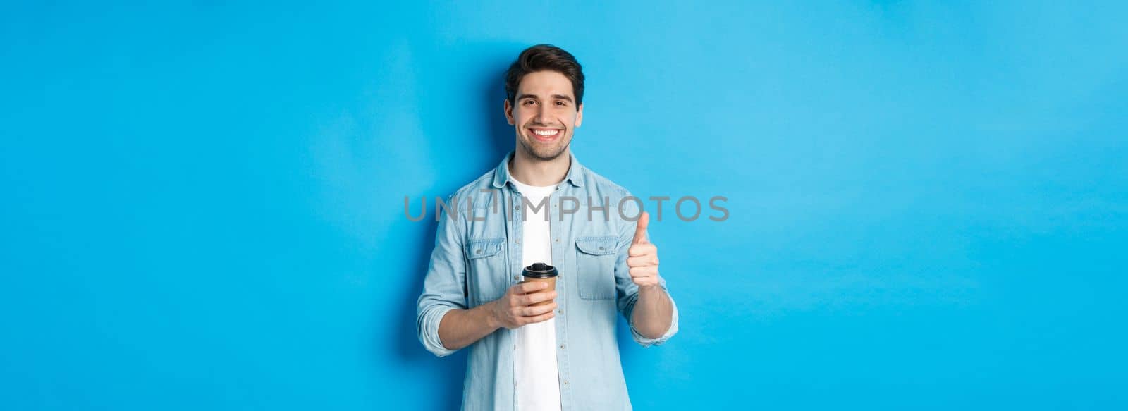 Handsome young man showing thumb-up and drinking coffee, recommending cafe takeaway, standing over blue background.