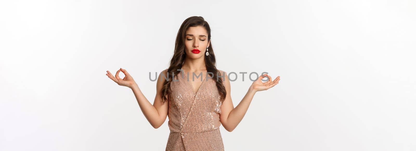 Christmas party and celebration concept. Calm and patient young woman in stylish dress meditating, feeling zen, standing over white background by Benzoix