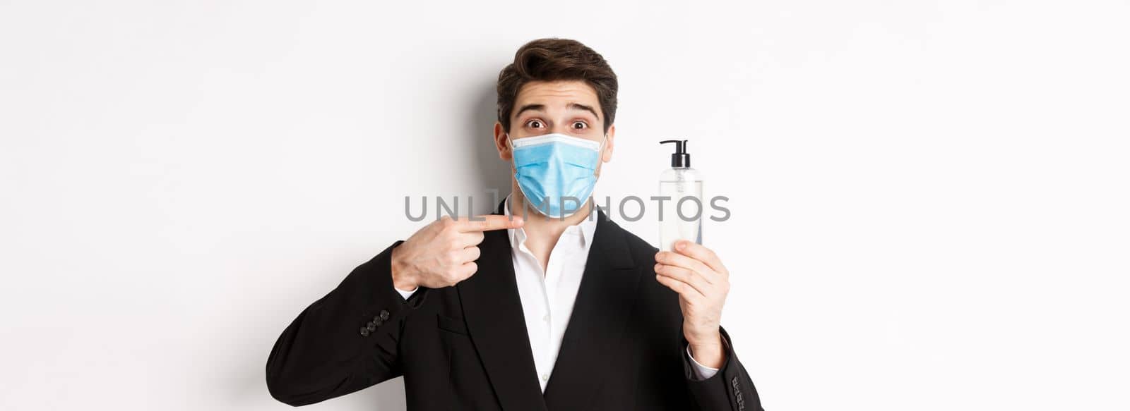 Concept of covid-19, business and social distancing. Close-up of handsome man in trendy suit and medical mask, recommending hand sanitizer, standing against white background by Benzoix