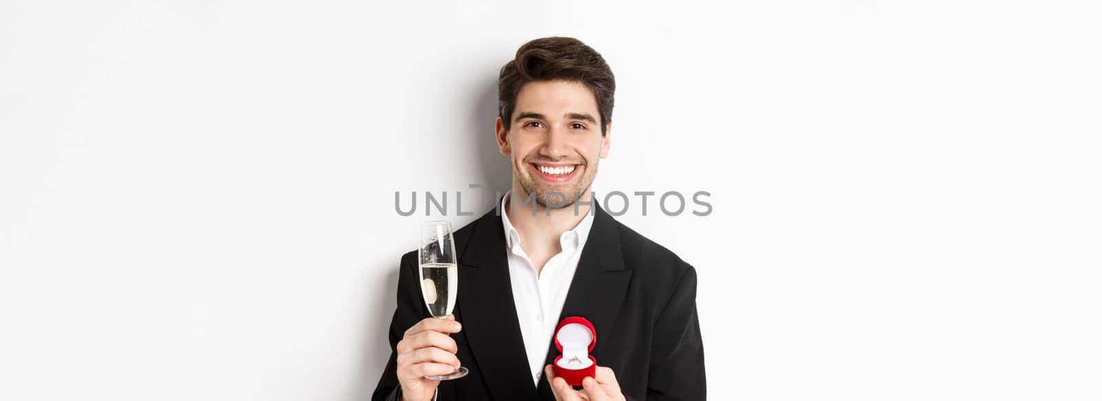 Close-up of handsome man in suit, making a proposal, giving engagement ring and raising glass of champagne, standing against white background.