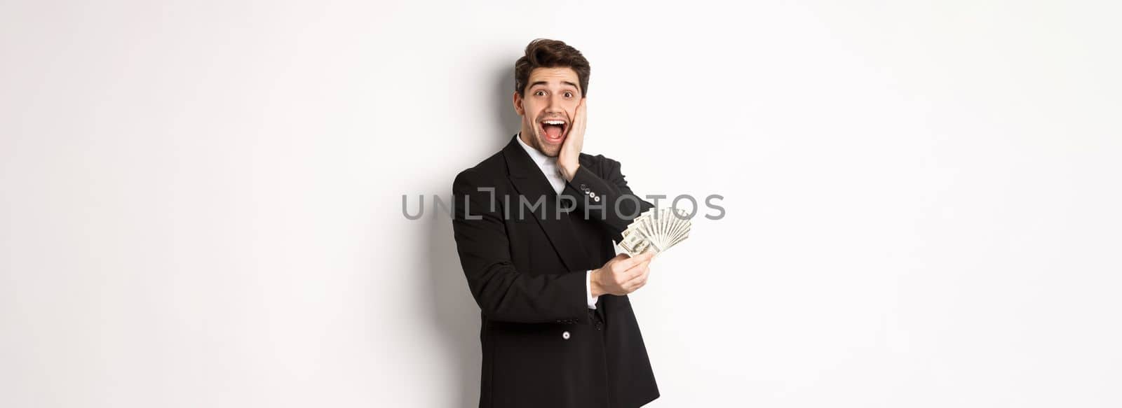 Image of rich and happy man in black suit, winning prize, holding money and looking excited at camera, standing over white background.