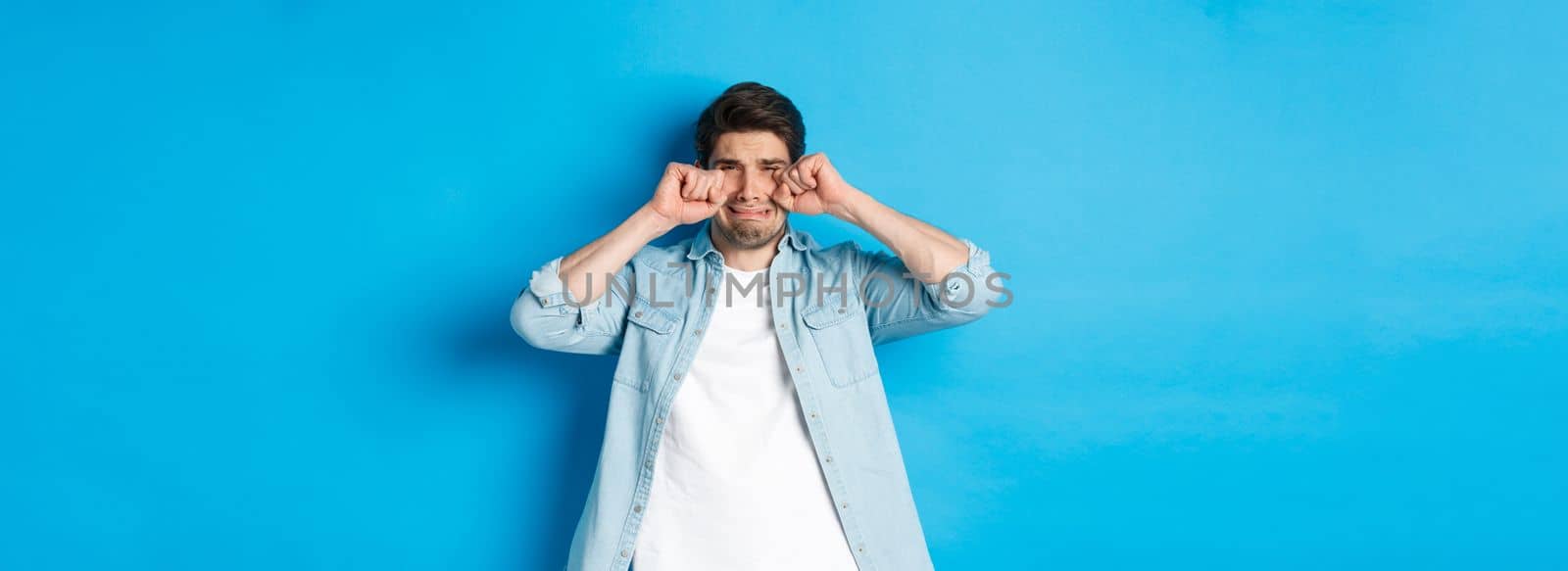 Sad timid man crying, wiping tears off face and sobbing, looking offended and upset, standing over blue background.