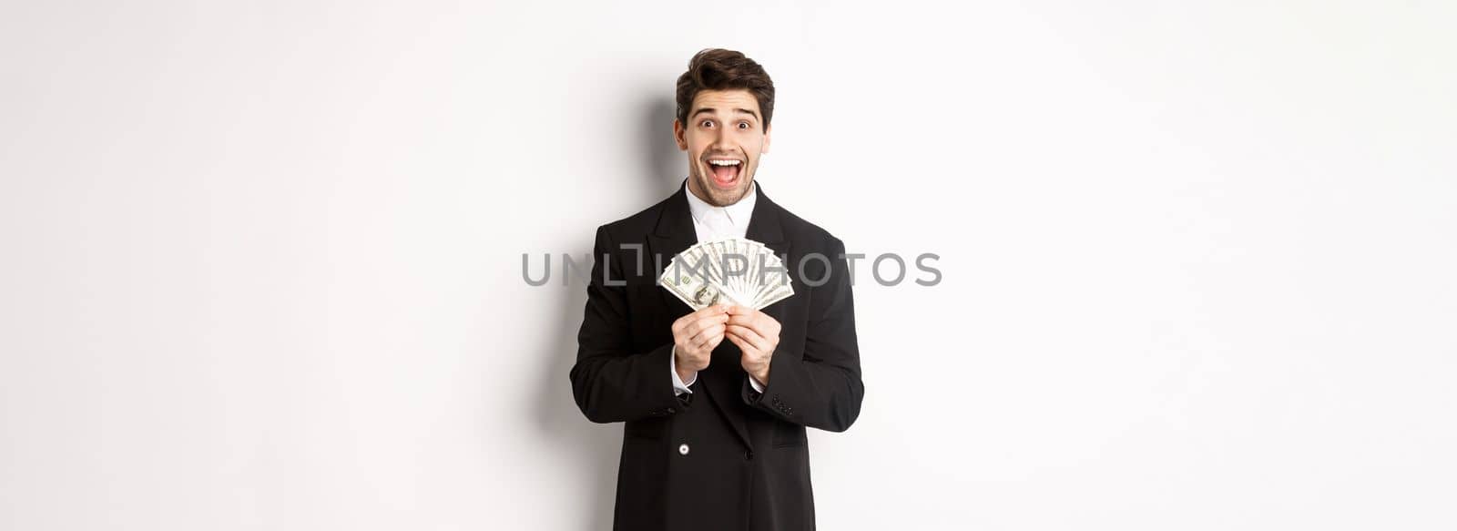Portrait of handsome successful businessman in trendy suit, showing money and smiling amazed, earn cash, standing over white background by Benzoix