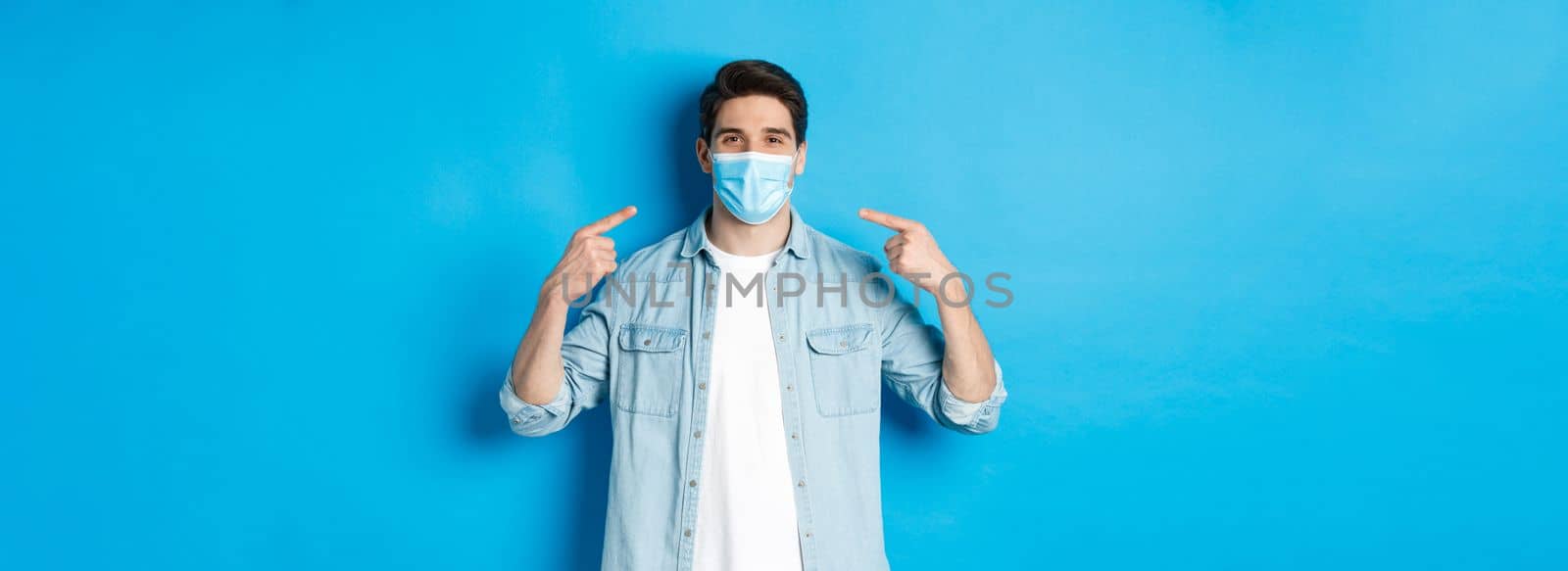 Concept of coronavirus, quarantine and social distancing. Handsome man pointing at medical mask and smiling, protection from virus spread during pandemic, blue background.