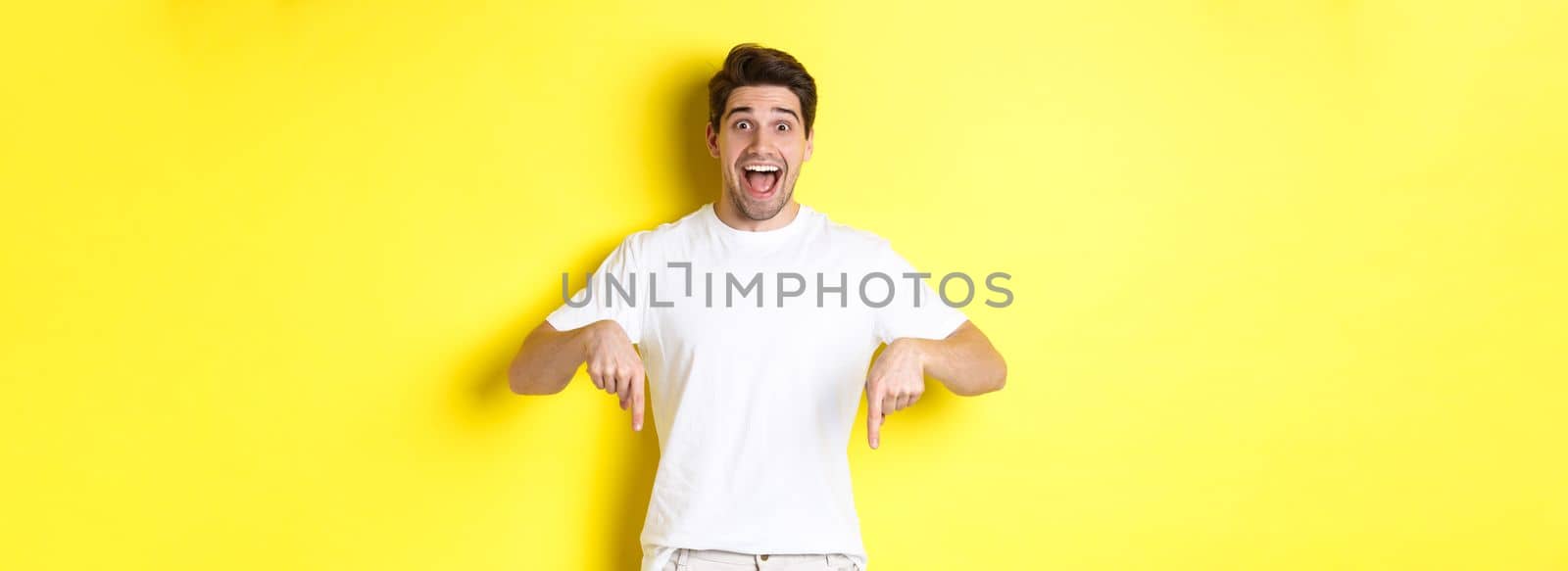 Amazed handsome man pointing fingers down, showing banner with excitement, standing over yellow background.