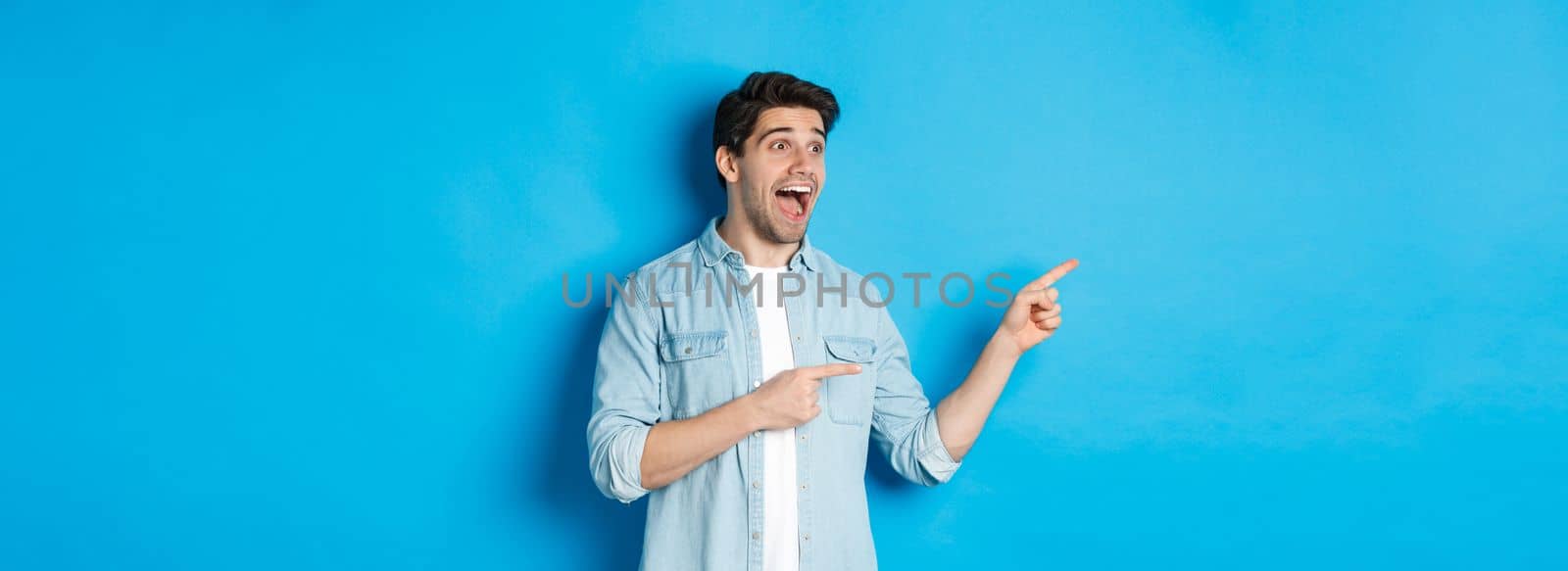 Portrait of handsome man looking and pointing fingers left with excitement, standing against blue background.
