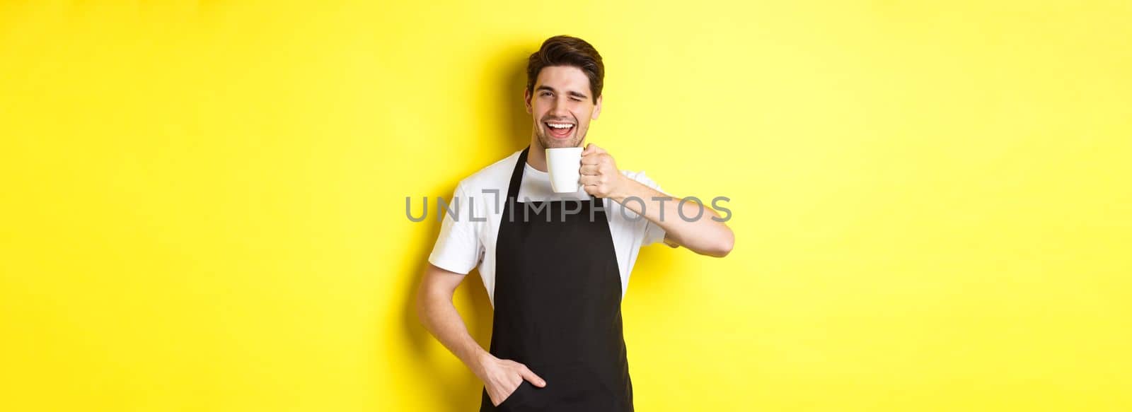 Handsome barista drinking cup of coffee and winking, inviting to cafe, standing over yellow background by Benzoix