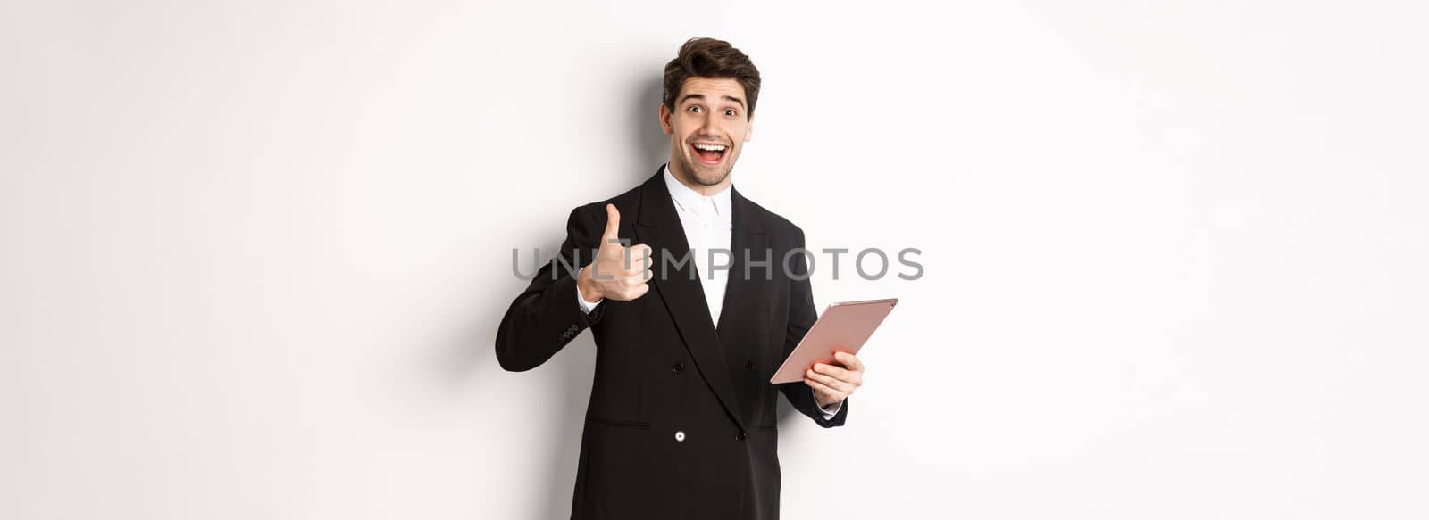 Portrait of handsome businessman in suit, showing thumb-up and making a compliment, praising something on digital tablet, standing over white background by Benzoix