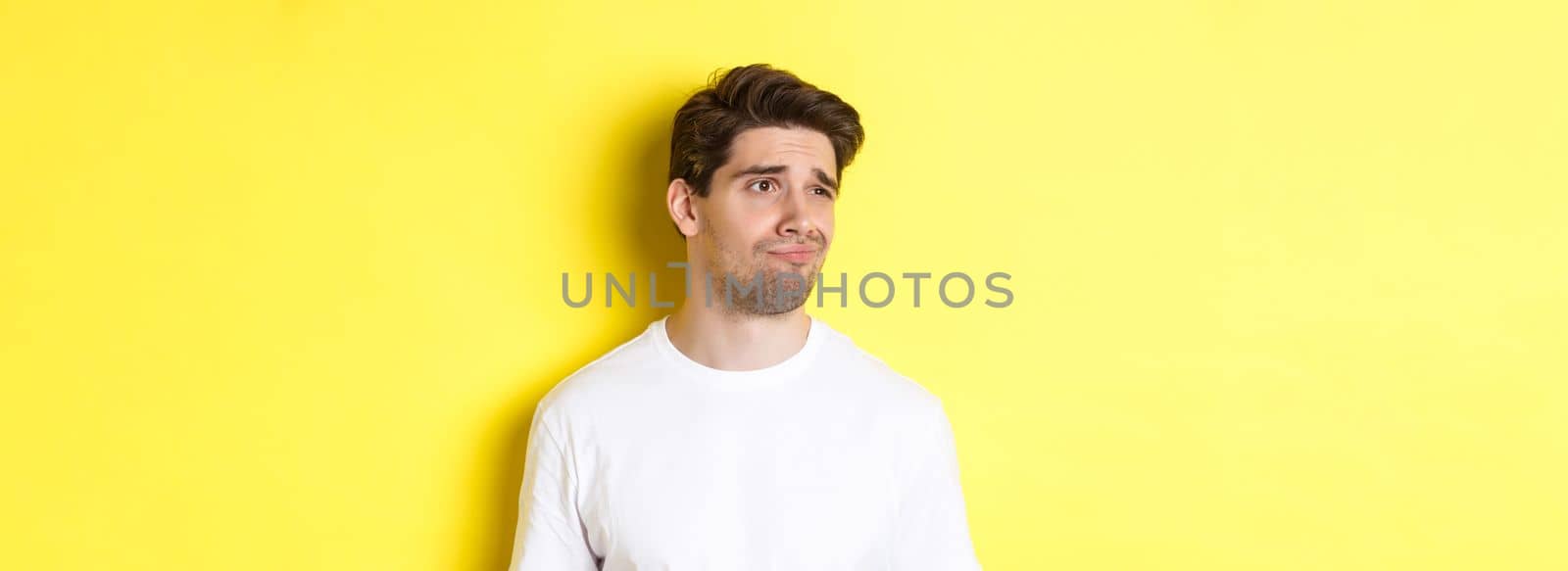 Reluctant guy in white t-shirt looking left, grimacing skeptical and displeased, standing over yellow background.