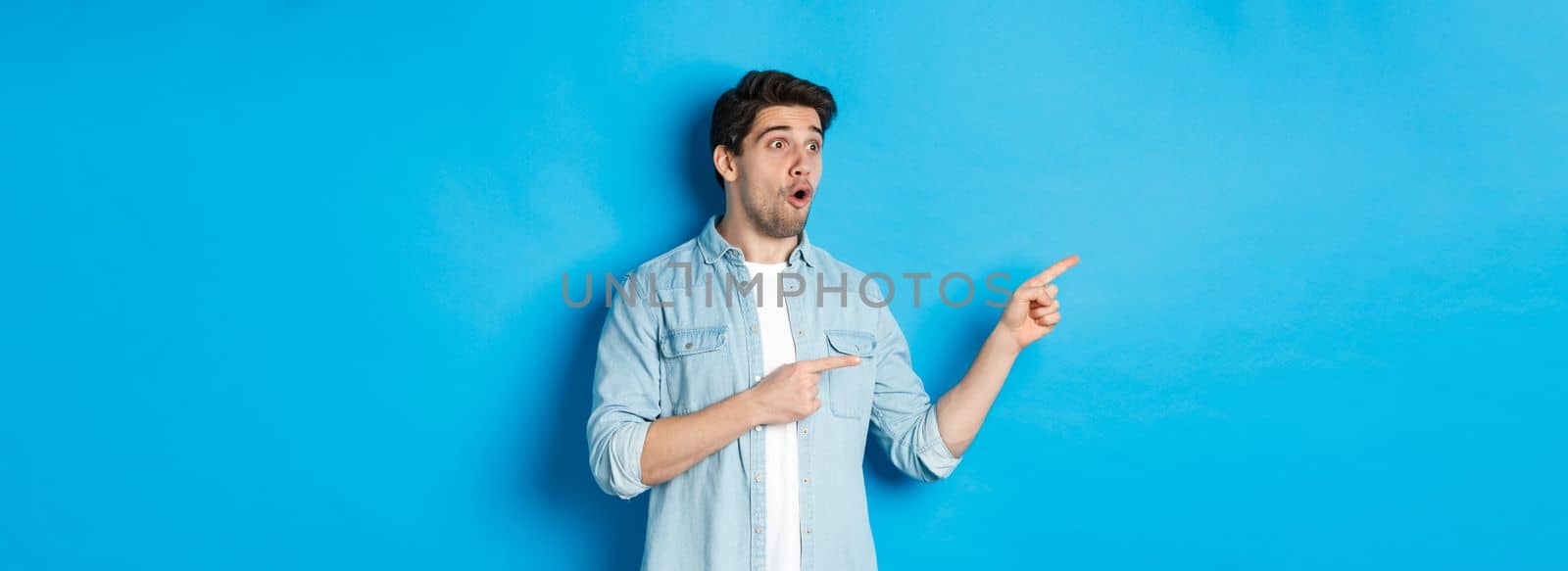 Surprised and amazed man looking at promotion, pointing fingers left at advertisement, standing over blue background by Benzoix