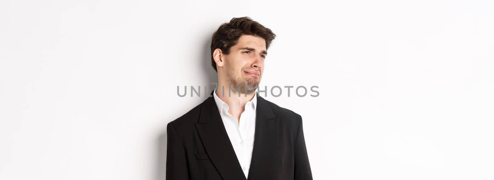 Close-up of disgusted young man in trendy suit, grimacing upset, looking left and standing against white background.