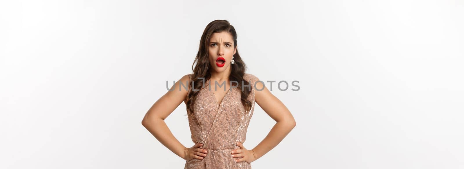 Displeased and shocked woman in party dress and makeup, frowning upset, gasping and looking at something disappointing, complaining, standing over white background.