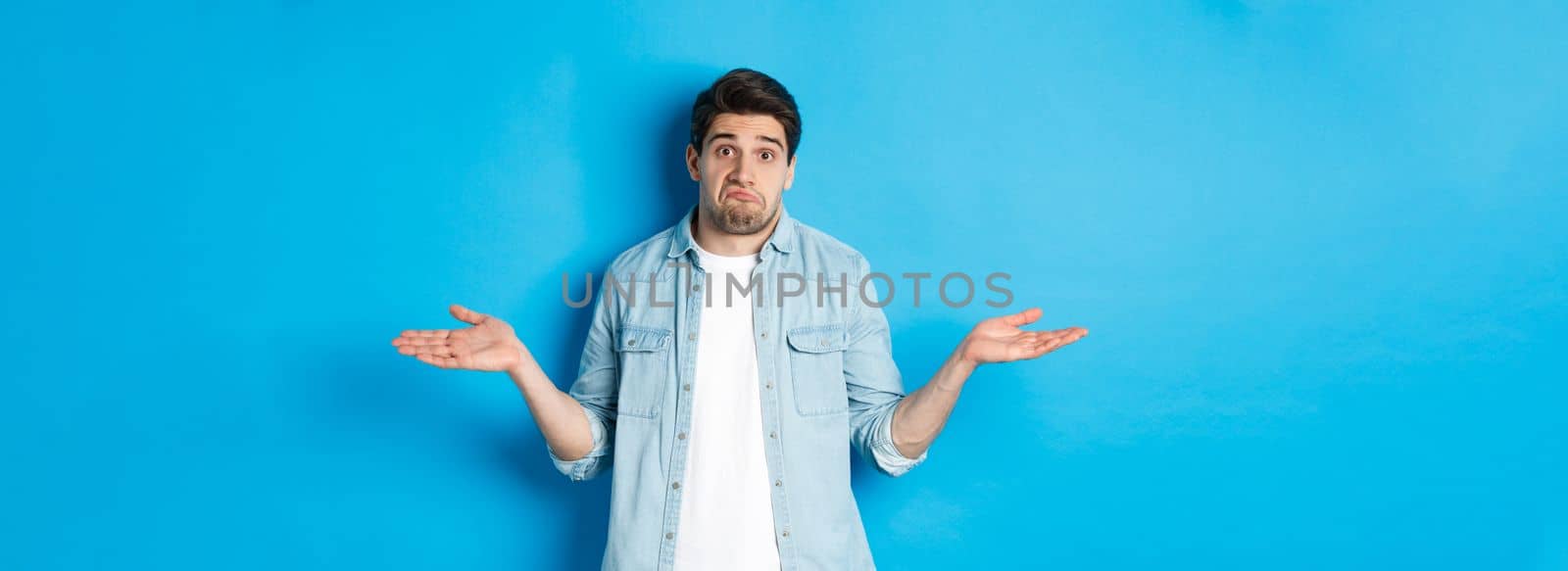 Confused young man in casual clothes shrugging, dont know anything, looking clueless, standing against blue background.