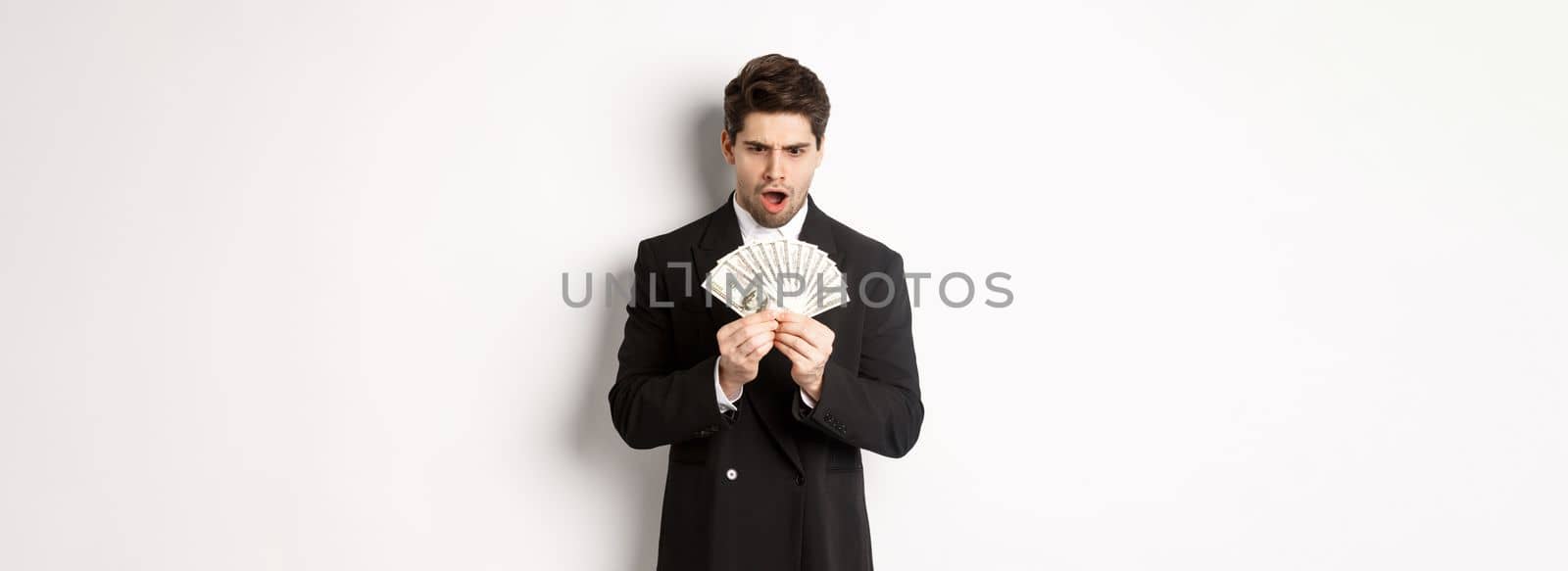 Portrait of handsome businessman in trendy suit, looking startled at money, won prize, standing over white background by Benzoix
