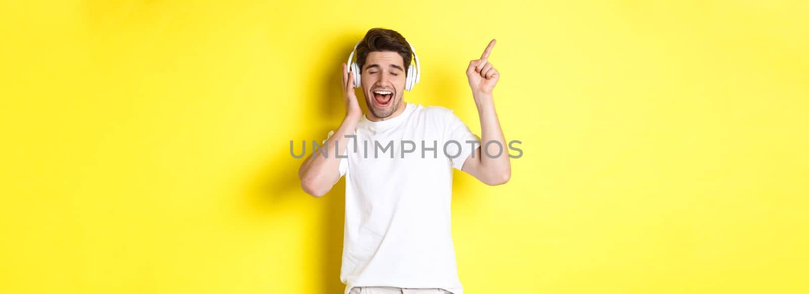 Happy man listening music in headphones, pointing finger at promo offer for black friday, standing over yellow background.