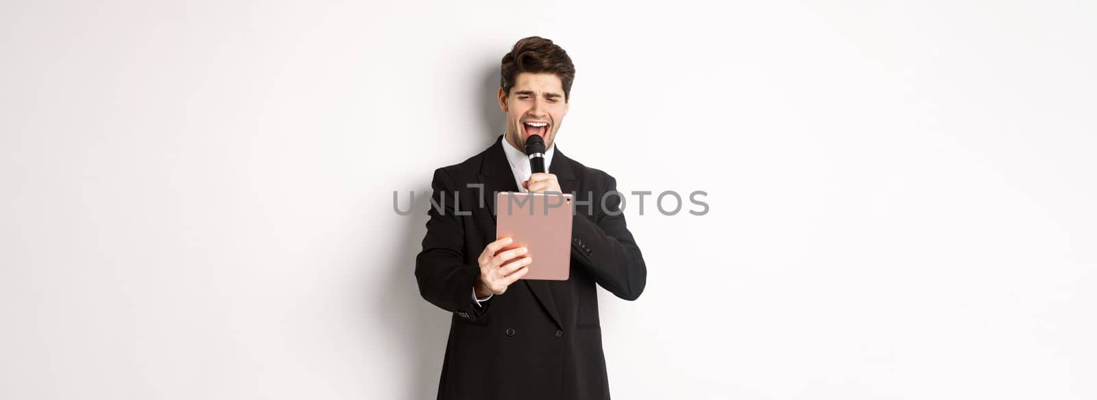 Image of handsome man in black suit, singing karaoke on digital tablet, holding microphone, standing over white background.