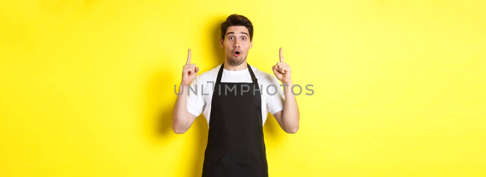 Surprised male waiter in black apron showing logo or advertisement, pointing fingers up at copy space, standing over yellow background by Benzoix