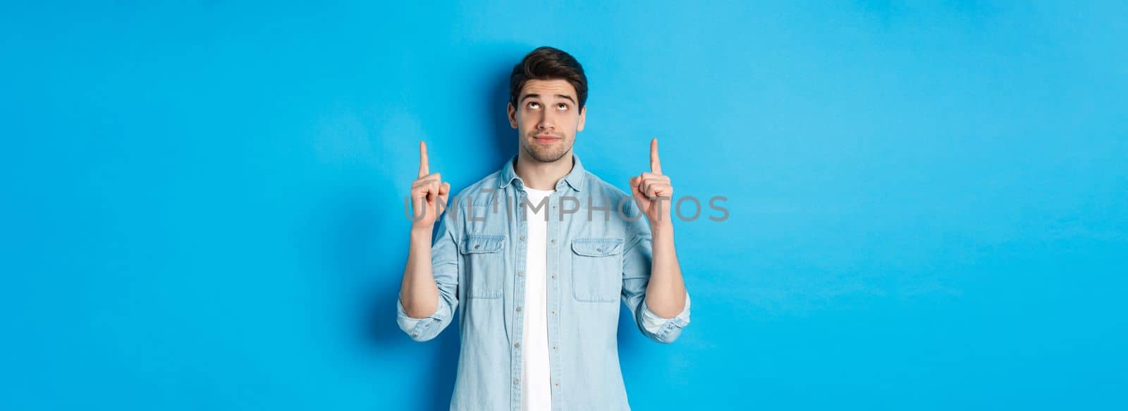 Portrait of displeased and skeptical male model pointing fingers up, looking at something unpleasant, standing against blue background by Benzoix
