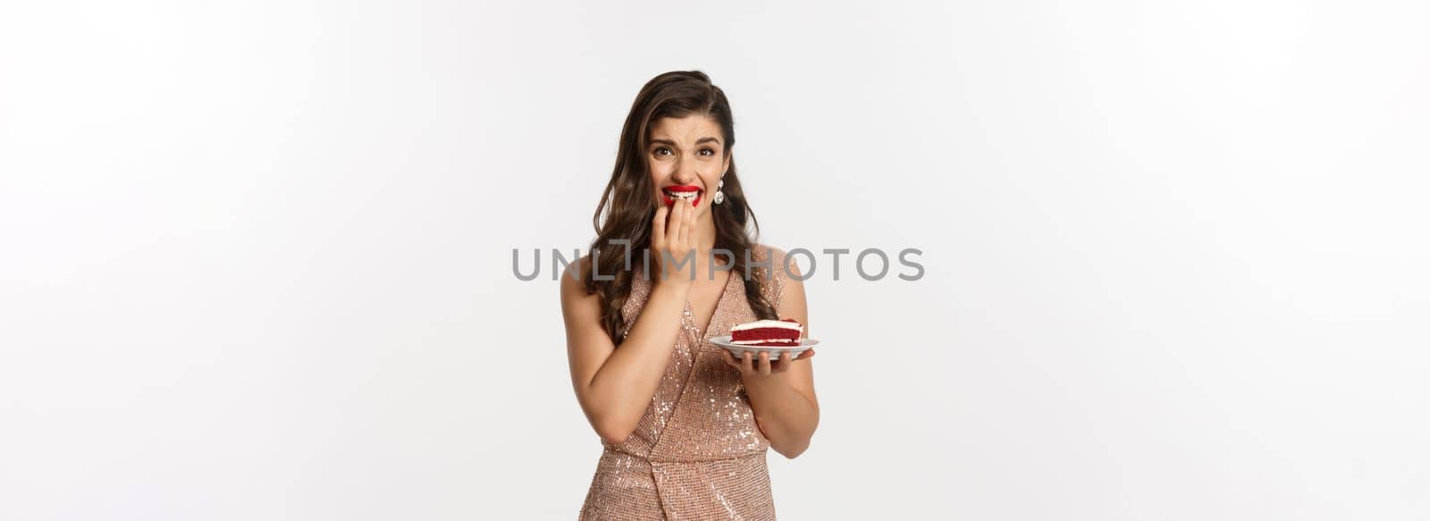 Image of young woman hesitating to eat cake on party, biting fingernails from temptation, standing over white background in elegant dress by Benzoix