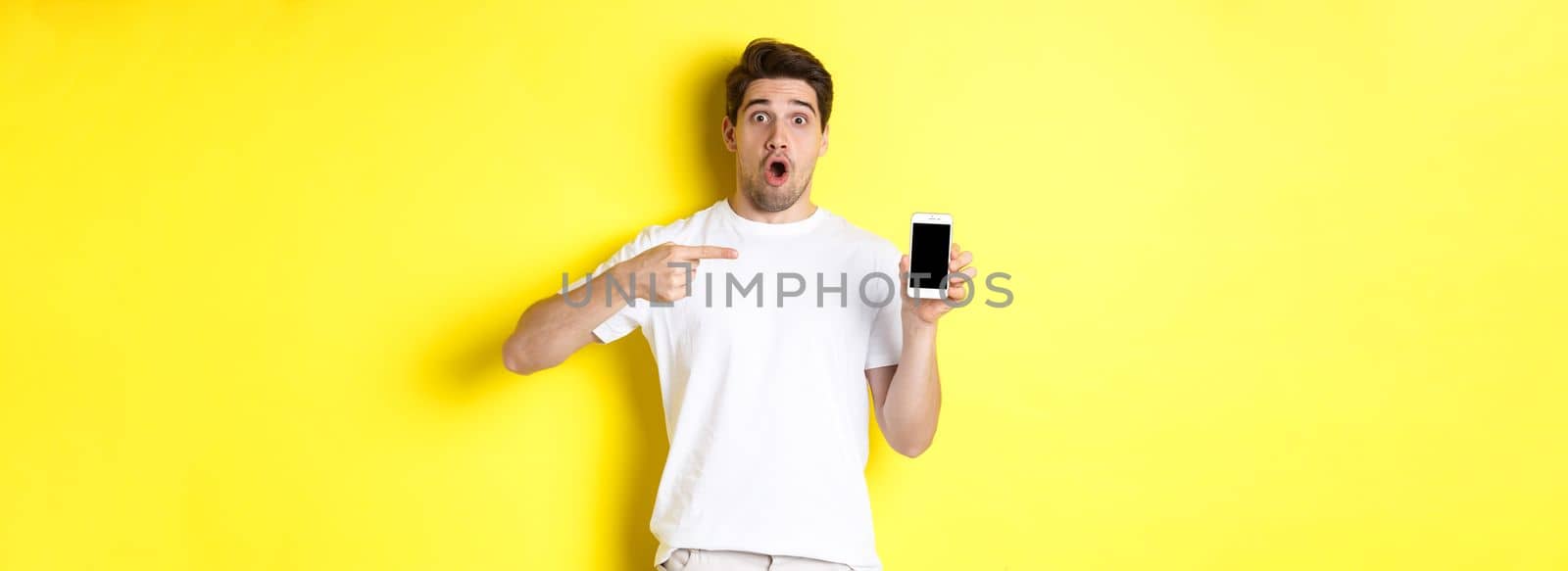 Impressed guy check out smartphone screen, pointing finger at display and gasping amazed, standing over yellow background by Benzoix