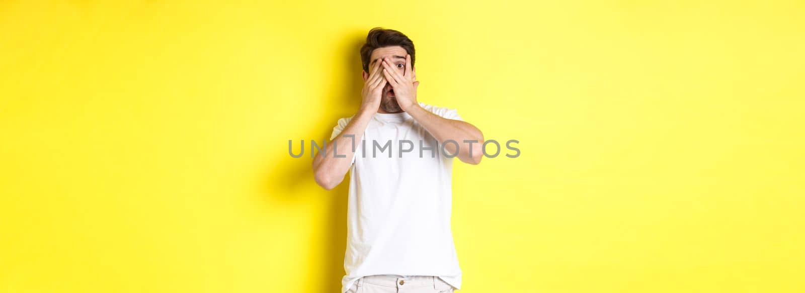 Embarrassed guy shut eyes but peeking through fingers at something awkward, standing over yellow background.