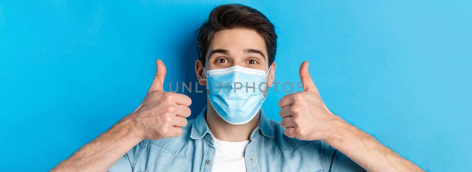 Concept of covid-19, pandemic and quarantine. Close-up of cheerful young man in medical mask smiling, showing thumbs up in approval, like and agree, standing over blue background by Benzoix