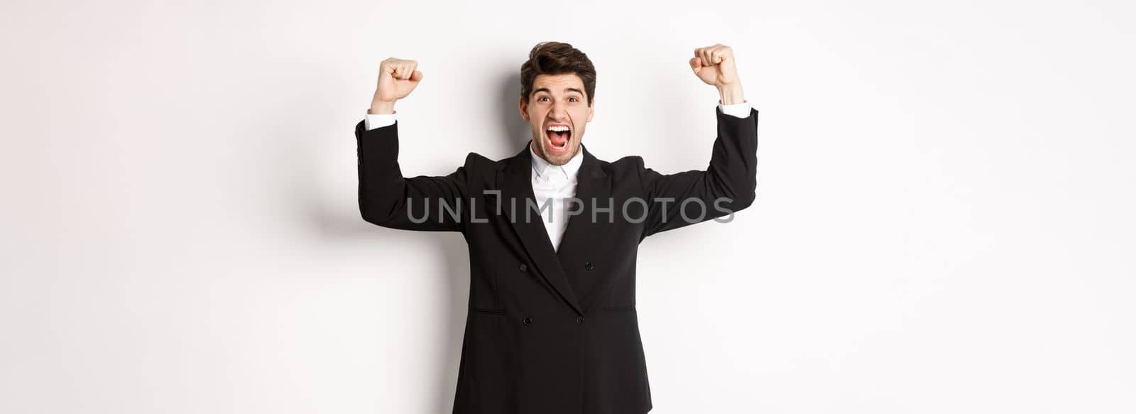 Successful handsome businessman triumphing, raising hands up and shouting yes, rejoicing over achievement, standing against white background.