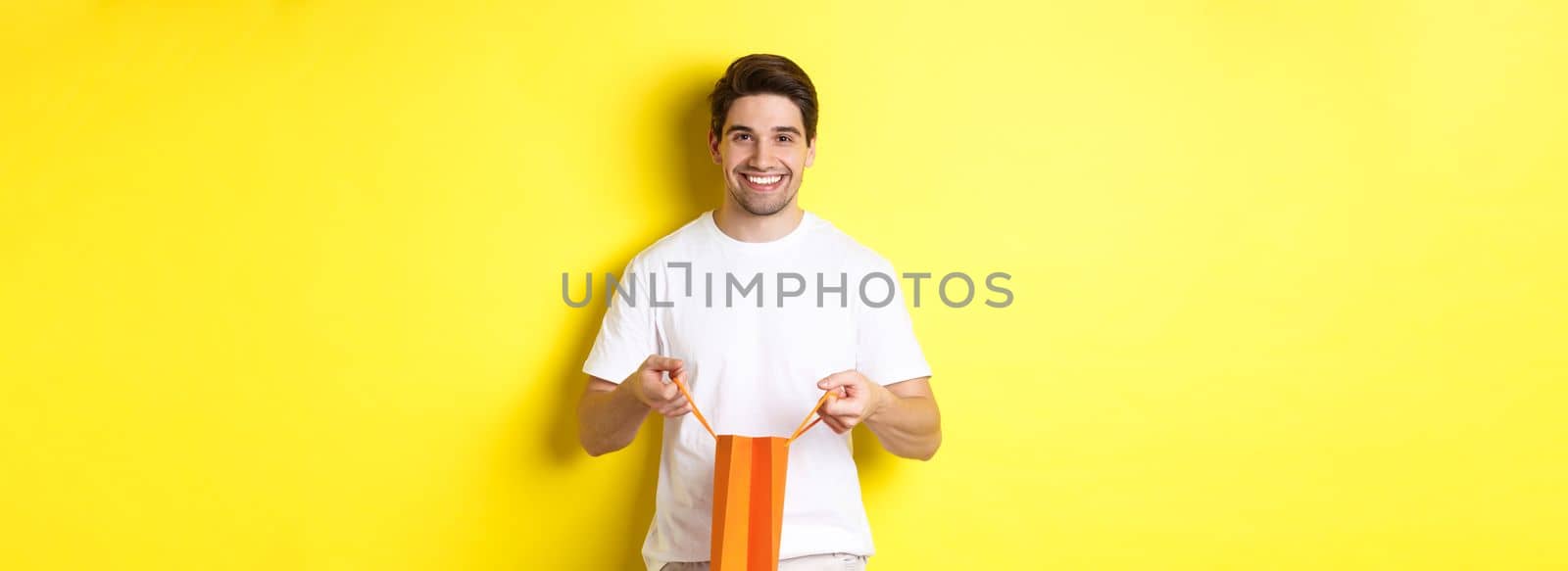 Happy young man open shopping bag with present, smiling at camera, standing against yellow background.