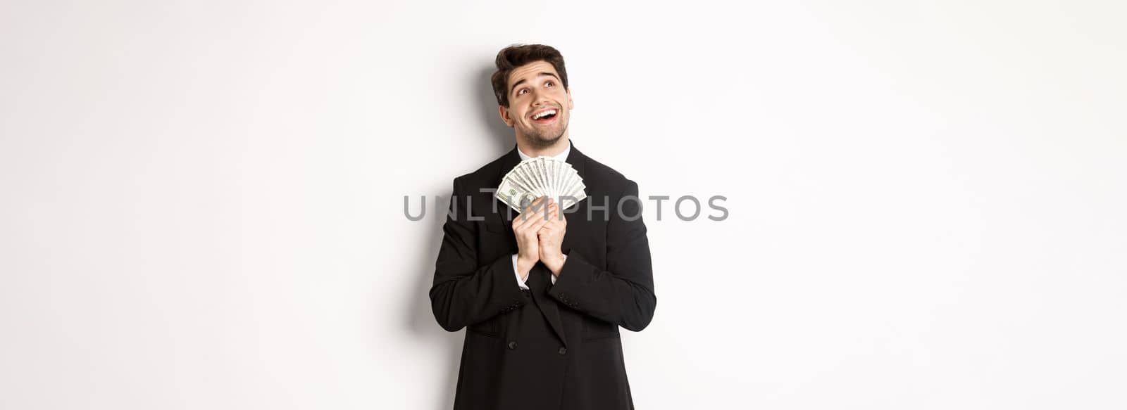 Image of handsome dreamy man in black suit, holding money and looking at upper left corner, thinking about shopping, standing over white background.