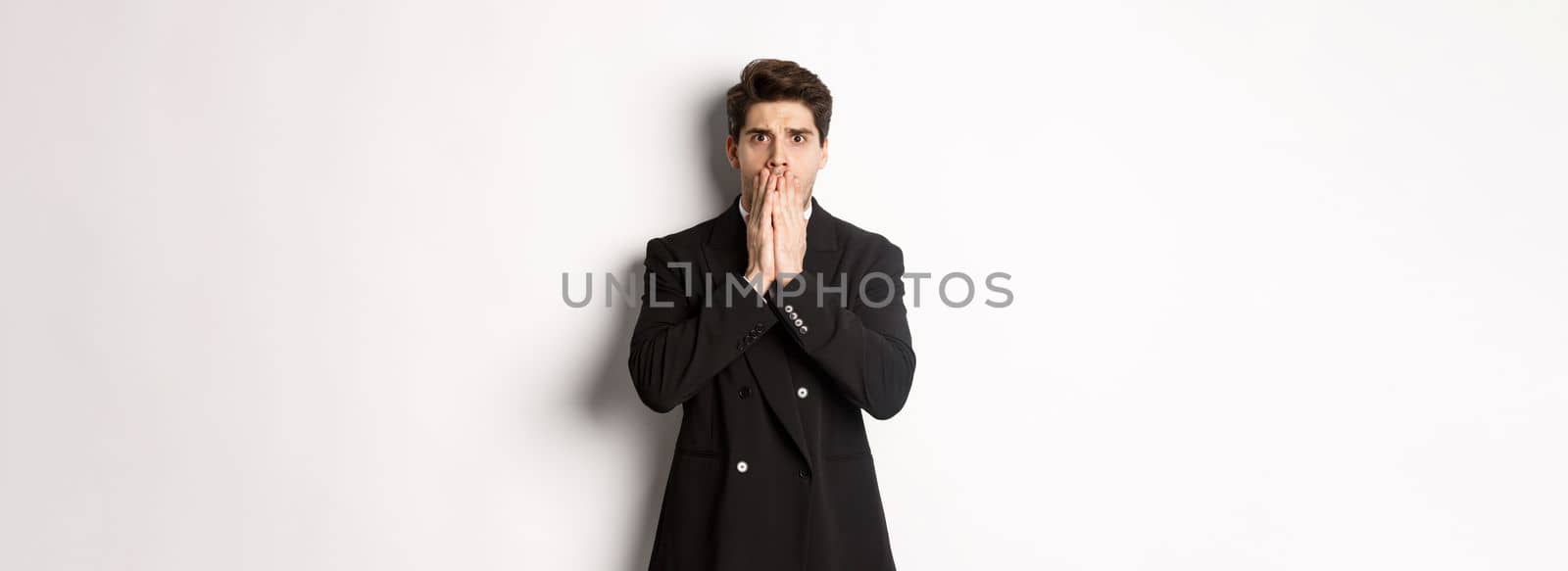 Portrait of shocked handsome businessman in suit, reacting to terrible situation, gasping and covering mouth with hands, standing startled against white background.