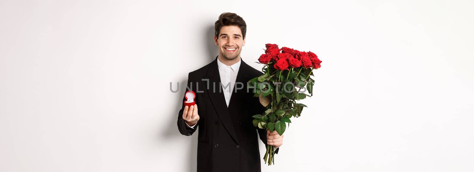 Handsome smiling man in black suit, holding roses and engagement ring, making a proposal to marry him, standing against white background by Benzoix