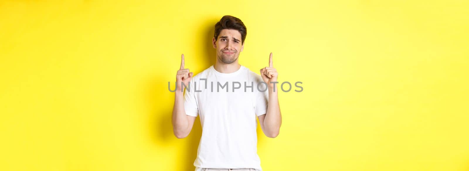 Unamused handsome guy frowning, pointing fingers up at something bad, standing skeptical against yellow background.