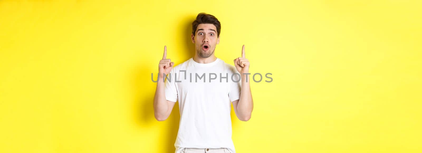 Surprised handsome guy in white t-shirt, pointing fingers up, interested about advertisement, standing against yellow background by Benzoix