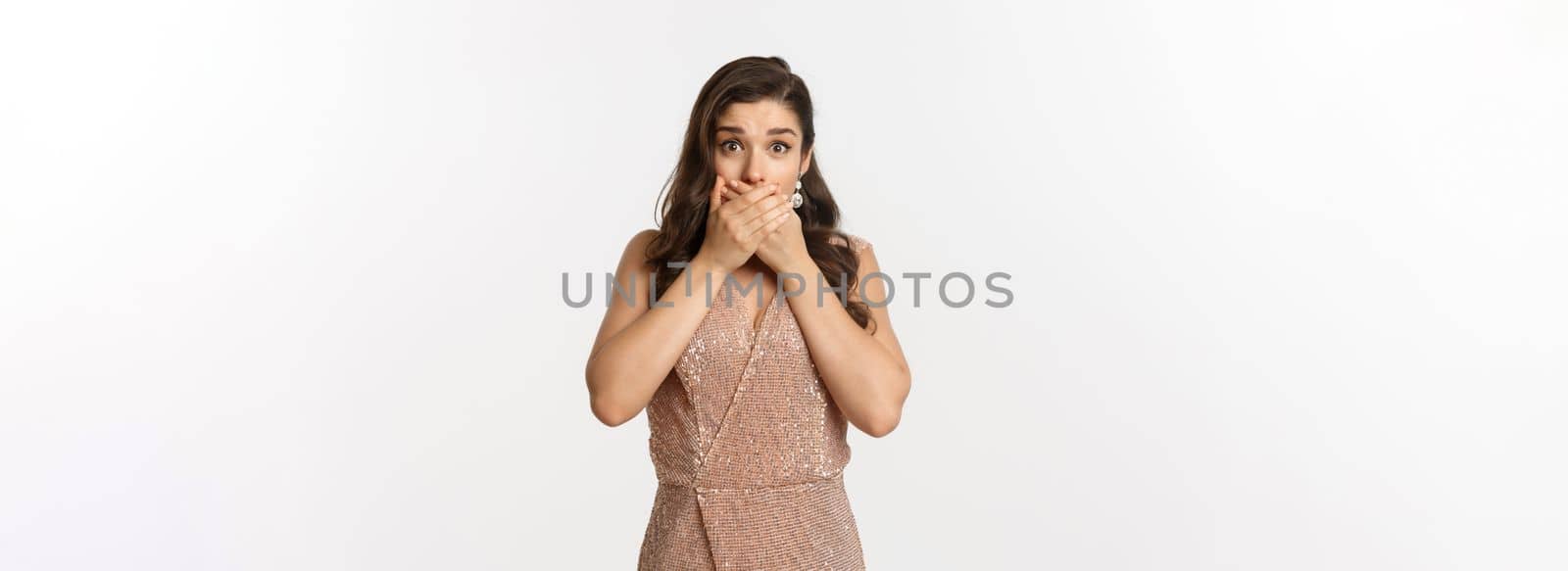 Concept of celebration, holidays and party. Startled young woman in glamour dress gasping and covering mouth with hands, looking shocked at camera, white background.