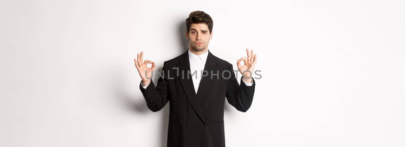 Concept of new year party, celebration and lifestyle. Portrait of confident good-looking man in black suit, showing okay sign and approve something, standing over white background.