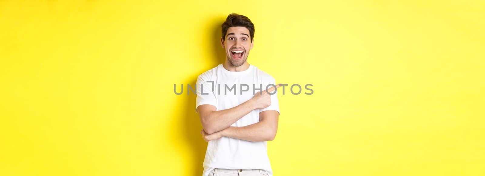 Happy man pointing finger left, showing advertisement on copy space, smiling amused, standing in white clothes against yellow background by Benzoix