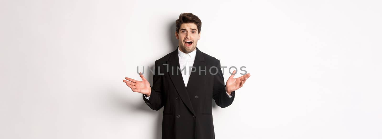 Portrait of confused and worried handsome man in suit, looking at something strange, spread hands sideways and standing puzzled against white background by Benzoix