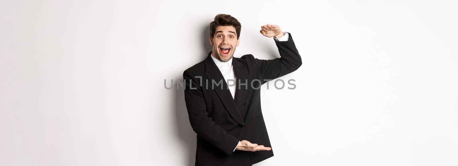 Portrait of excited handsome man in suit, shaping big object on copy space and smiling amazed, holding something, standing over white background.