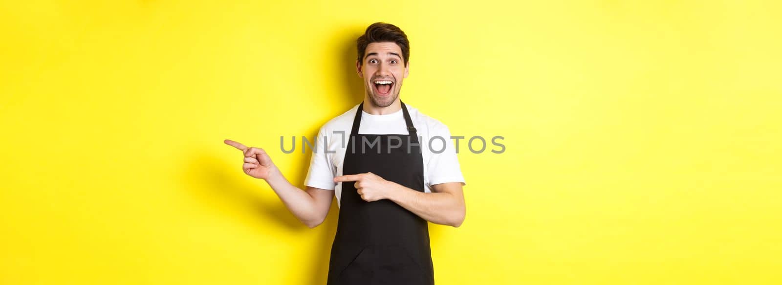 Waiter in black apron pointing fingers right, showing advertisement and smiling excited, standing against yellow background.