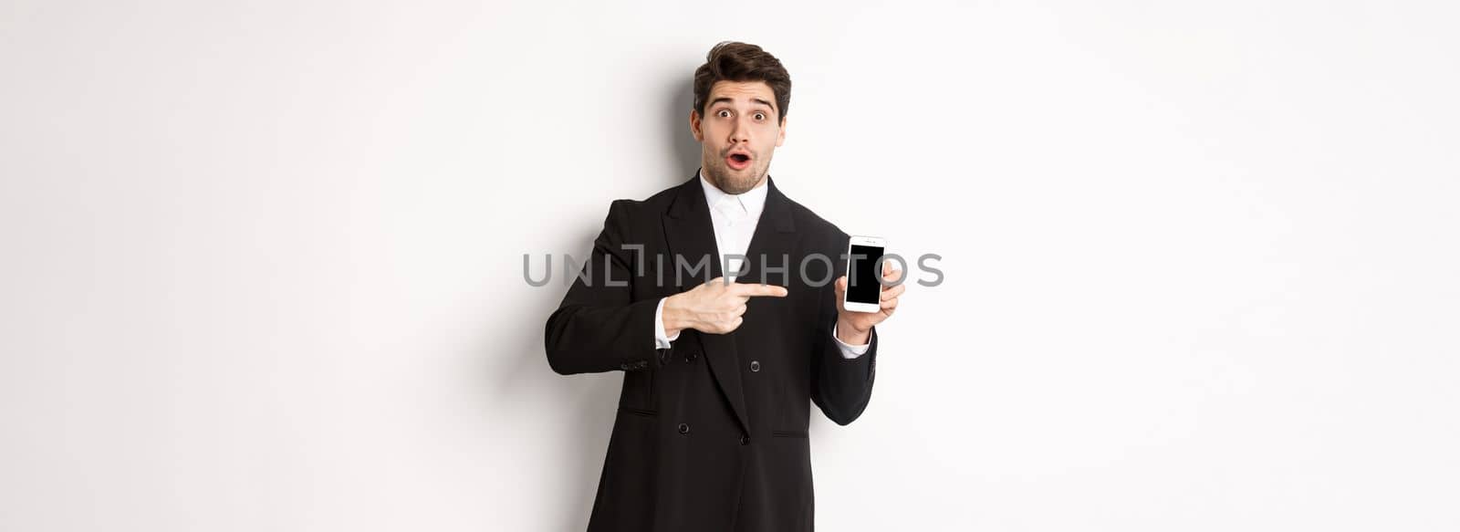 Portrait of attractive businessman in black suit, looking surprised and pointing finger at smartphone sreen, standing over white background by Benzoix