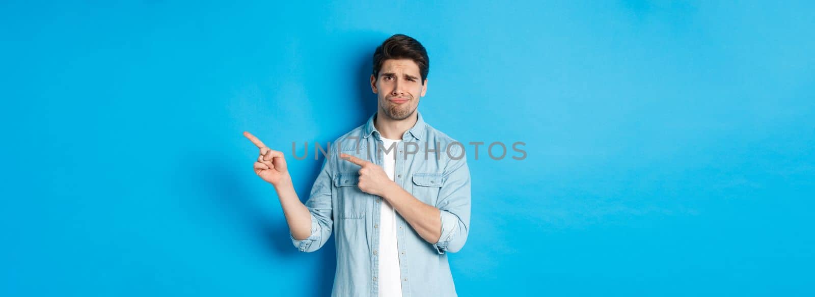 Portrait of skeptical adult guy pointing fingers right and smirking, exress disappointment and doubt, standing against blue background.