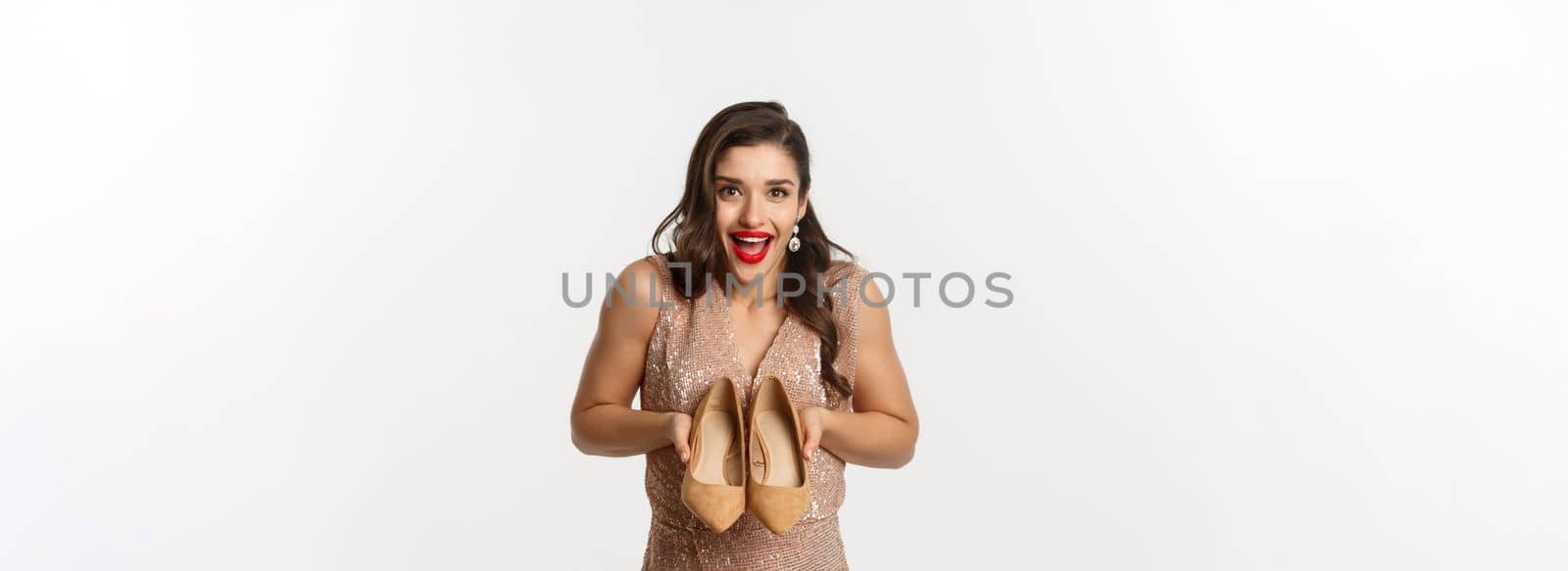 Party and celebration concept. Excited woman showing new pair of heels and smiling, wearing elegant dress, standing over white background.