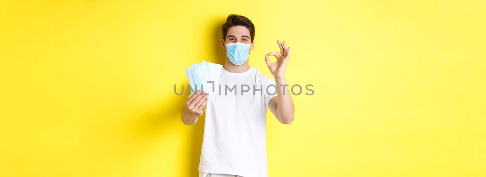 Concept of covid-19, quarantine and preventive measures. Satisfied man showing okay sign and giving medical masks, standing over yellow background by Benzoix