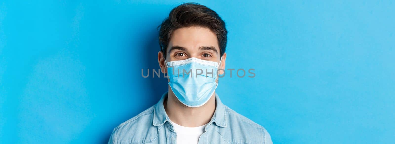 Concept of covid-19, pandemic and quarantine. Close-up of happy guy in medical mask looking at camera, standing over blue background.