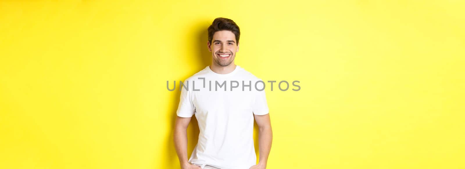 Young handsome man smiling at camera, holding hands in pockets, standing against yellow background.