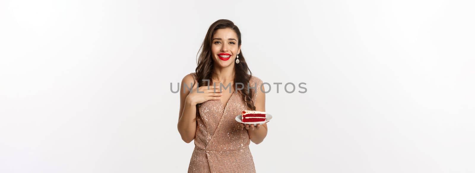 Party and celebration concept. Attractive slim woman in elegant dress holding piece of cake and smiling, standing over white background.