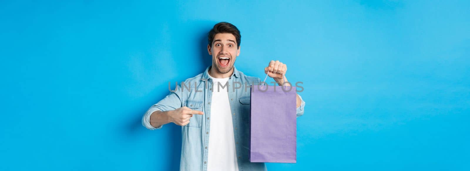 Concept of shopping, holidays and lifestyle. Excited guy pointing finger at paper bag and looking amazed, recommending store, announcing discounts, blue background.