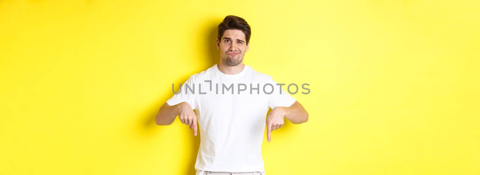 Young unhappy guy grimacing, pointing fingers down at advertisement, disappointed in product, standing over yellow background by Benzoix