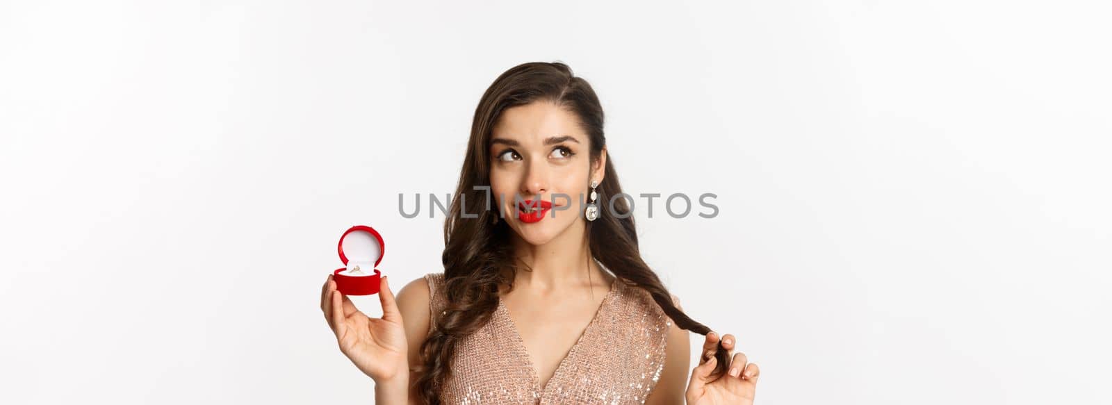 Close-up of coquettish beautiful woman thinking about marriage, holding engagement ring and looking at upper left corner, smiling silly, white background by Benzoix