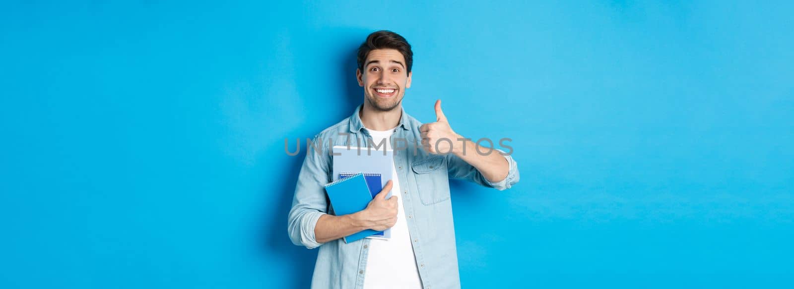 Happy young male model holding notebooks and showing thumb-up pleased, smiling and recommending courses, standing over blue background.